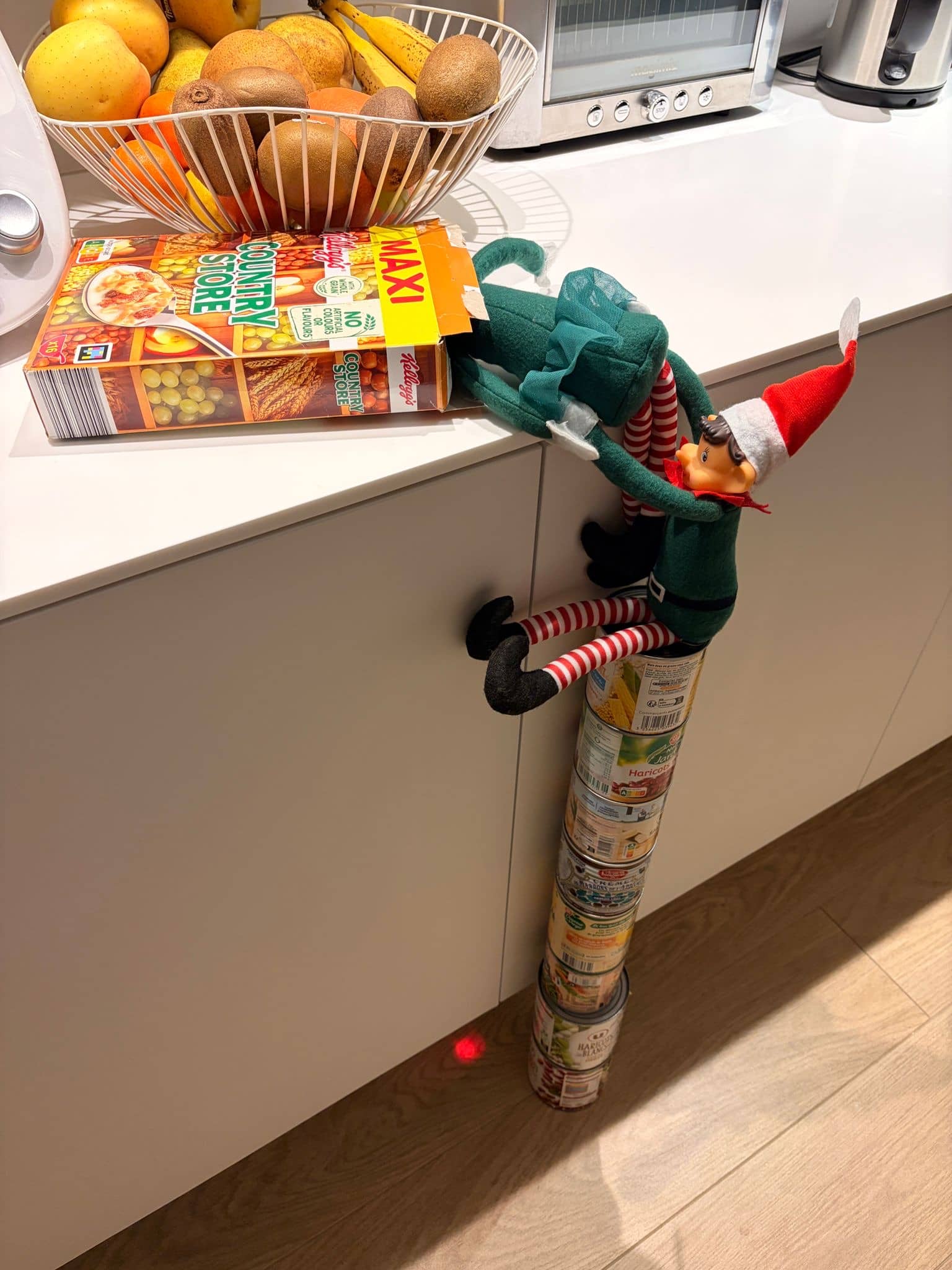 Lutin farceur en tenue de Noël grimpant sur des boîtes de conserve pour atteindre une boîte de céréales sur un plan de travail
