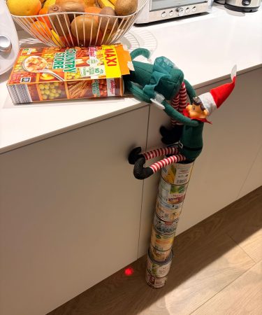 Lutin farceur en tenue de Noël grimpant sur des boîtes de conserve pour atteindre une boîte de céréales sur un plan de travail