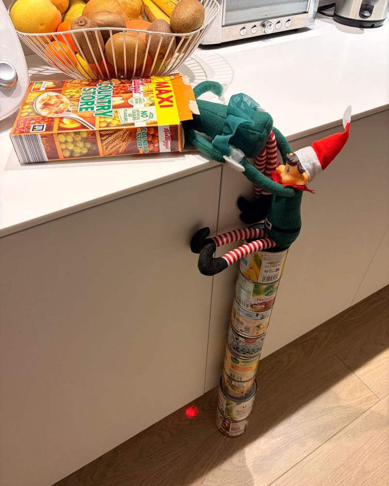 Lutin farceur en tenue de Noël grimpant sur des boîtes de conserve pour atteindre une boîte de céréales sur un plan de travail